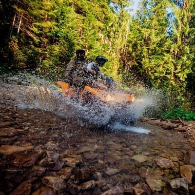 Двоє людей на квадроциклі їдуть річкою посеред лісу в Карпатах, бризки води на фоні природи Яремче